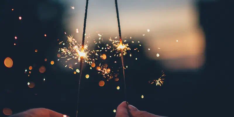 Two hands each holding a lit sparkler at dusk, sparks and soft orange lights against a dark blurred background.